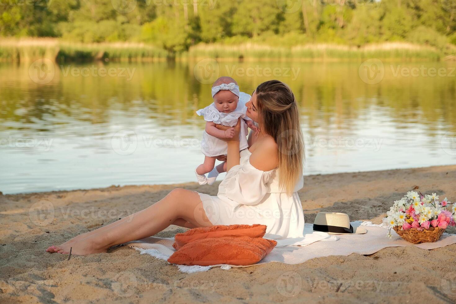 a beautiful girl in a white dress with a newborn baby is sitting on the beach, on the sand. photo