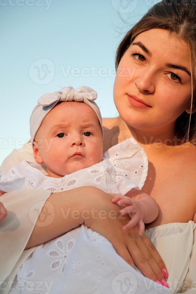 a beautiful girl in a white dress with a newborn baby is sitting on the beach, on the sand. photo