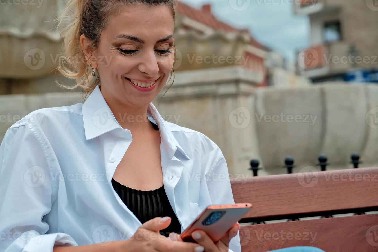 a beautiful girl sits on a bench and holds a phone in her hands. the girl orders food through the phone. the girl is talking on the phone laughing , happy , surprised . photo