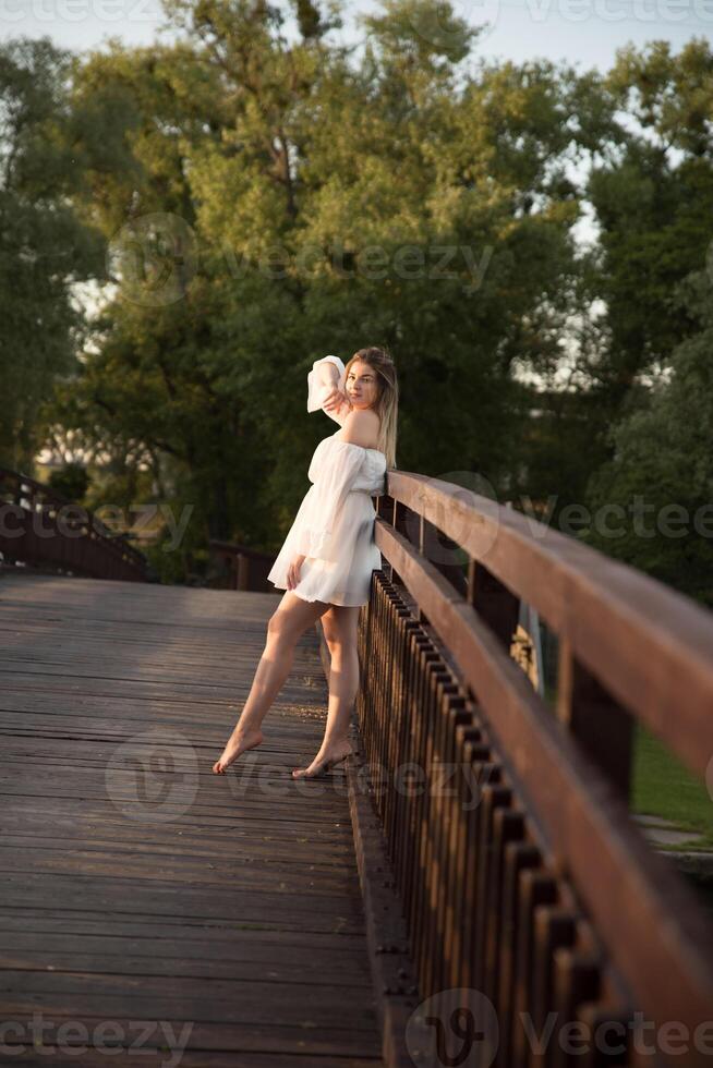 a beautiful girl in a white dress stands on a wooden bridge. photo