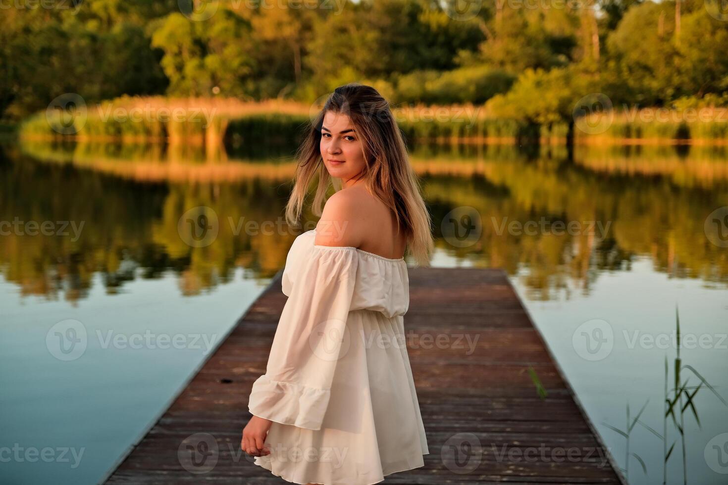 a beautiful girl is standing on the pier. in the background there is a river, a lake. photo