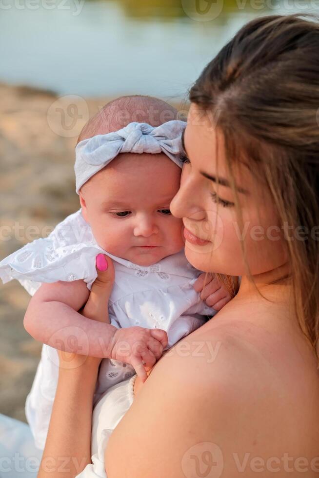 a beautiful girl in a white dress with a newborn baby is sitting on the beach, on the sand. photo