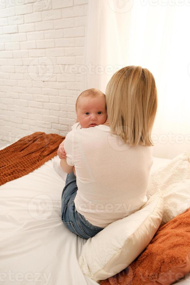 mom holds a newborn baby in her arms. in the background the sun shines from the window. photo