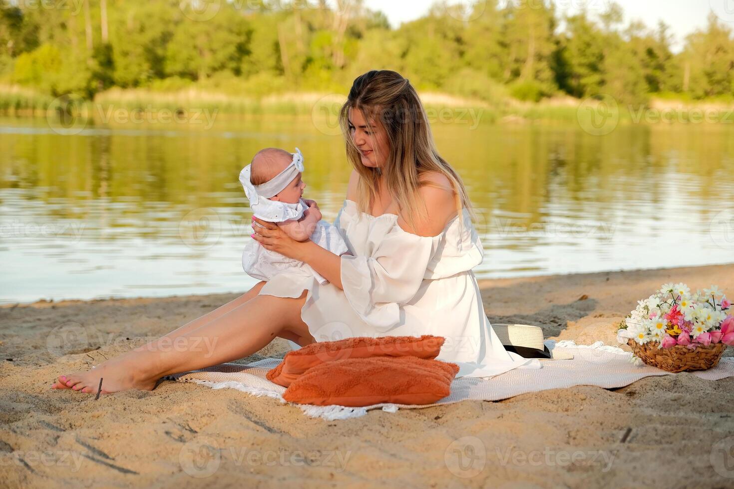 a beautiful girl in a white dress with a newborn baby is sitting on the beach, on the sand. photo