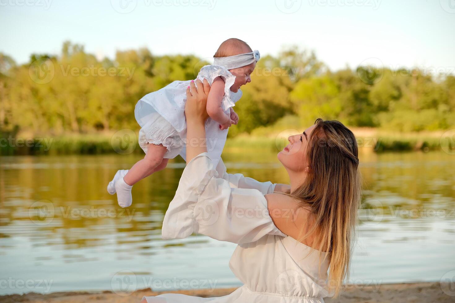 a beautiful girl in a white dress with a newborn baby is sitting on the beach, on the sand. photo