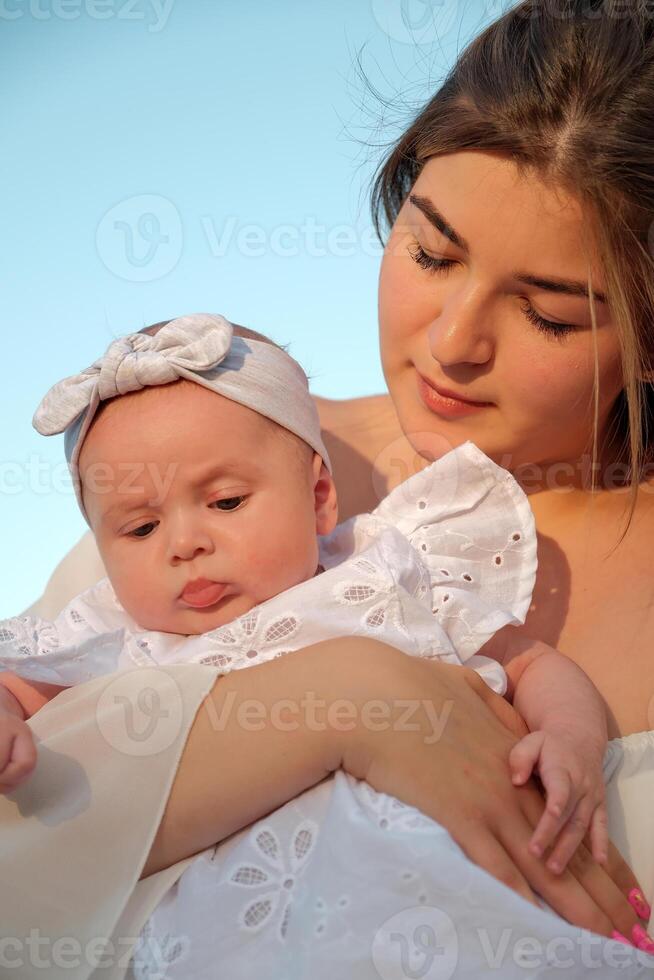 a beautiful girl in a white dress with a newborn baby is sitting on the beach, on the sand. photo