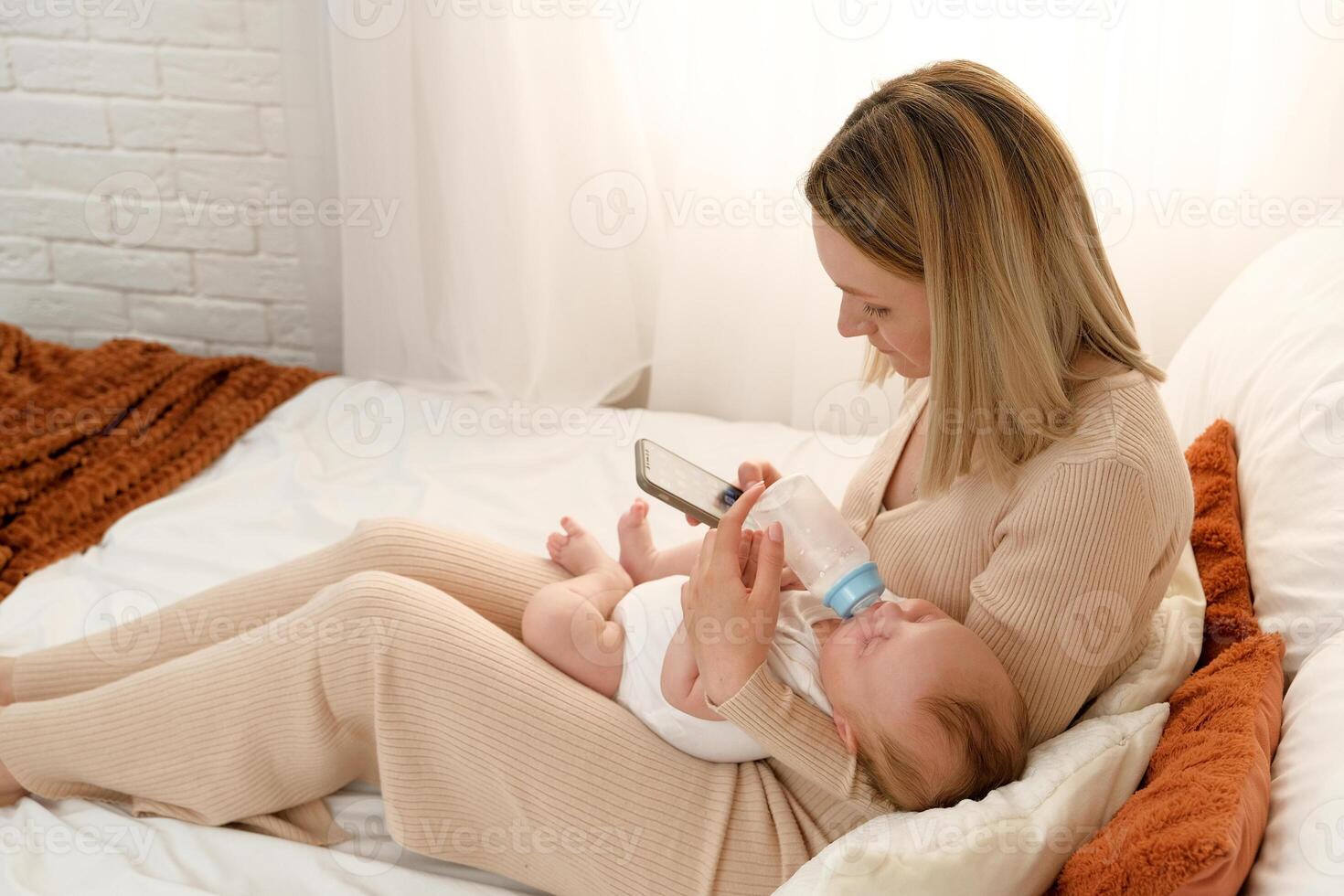 mom feeds a newborn baby with bottles of milk. mom holds the phone while feeding the baby. photo