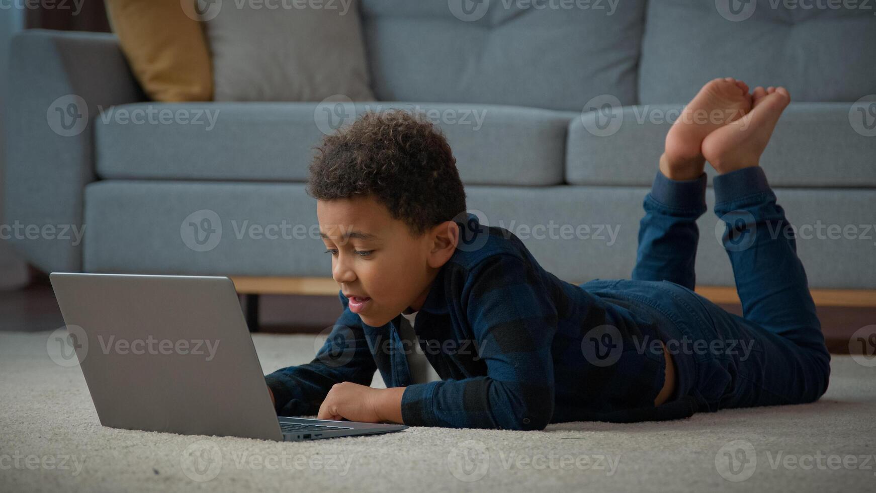 African American kid boy ethnic school pupil studying online from home watching web class lesson call e-learning. Little child son with laptop computer play game lying on floor in living room photo