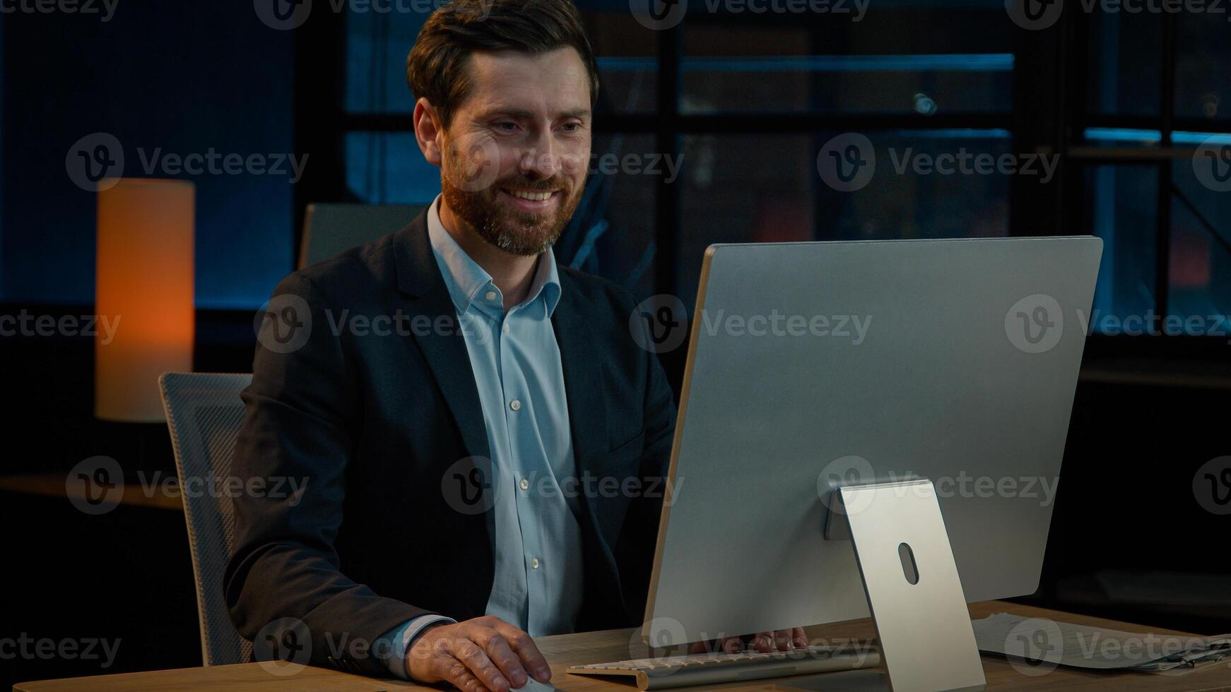 Middle-aged 40s businessman sit in night office company evening online job on computer business project startup. Caucasian adult man employer entrepreneur manager smiling satisfied smile good result photo