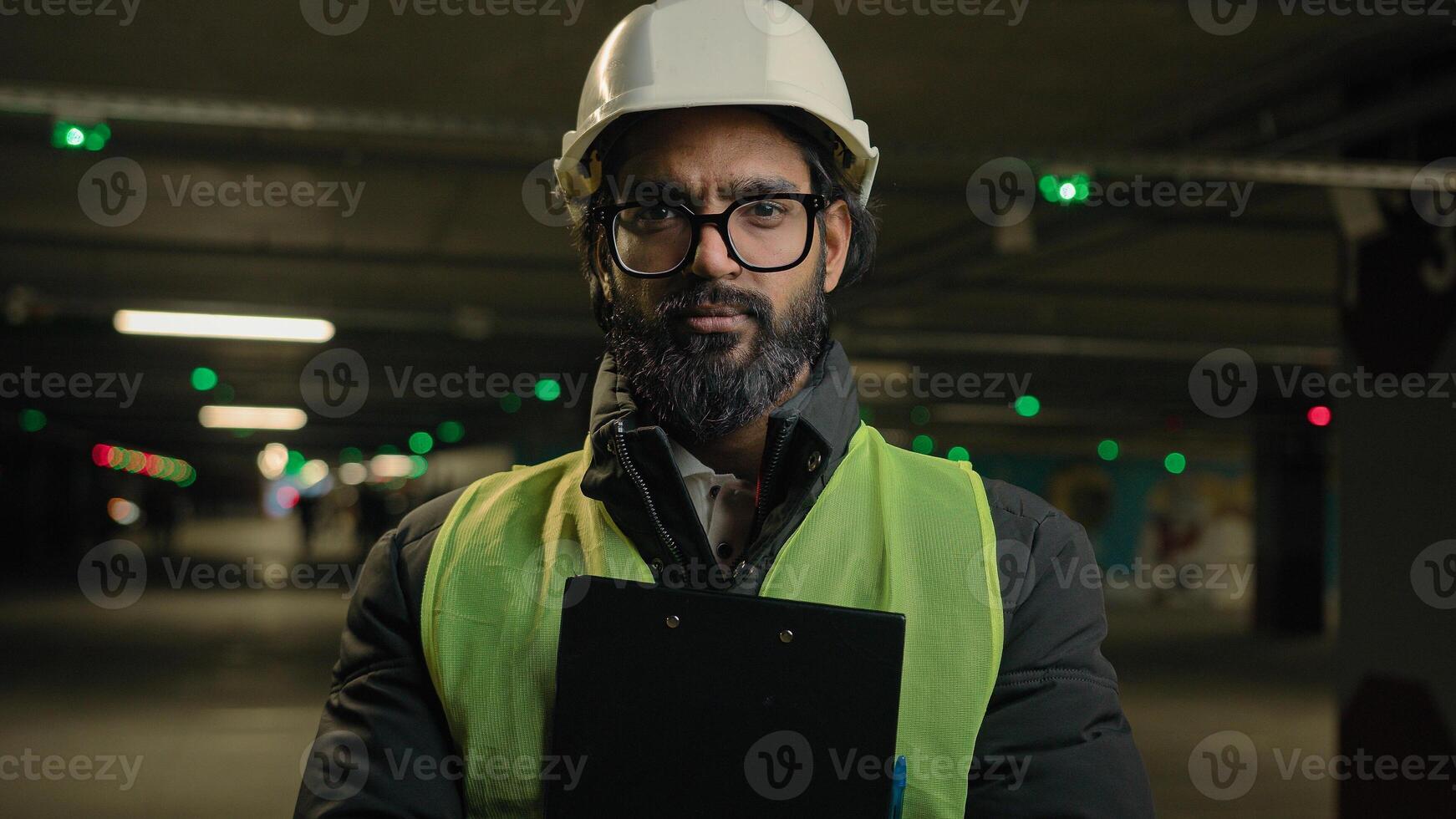 Portrait Indian man worker contractor mechanic guy in helmet and glasses posing with clipboard folder indoors parking factory warehouse manufacture. Arabian male builder engineer fitter in hard hat photo
