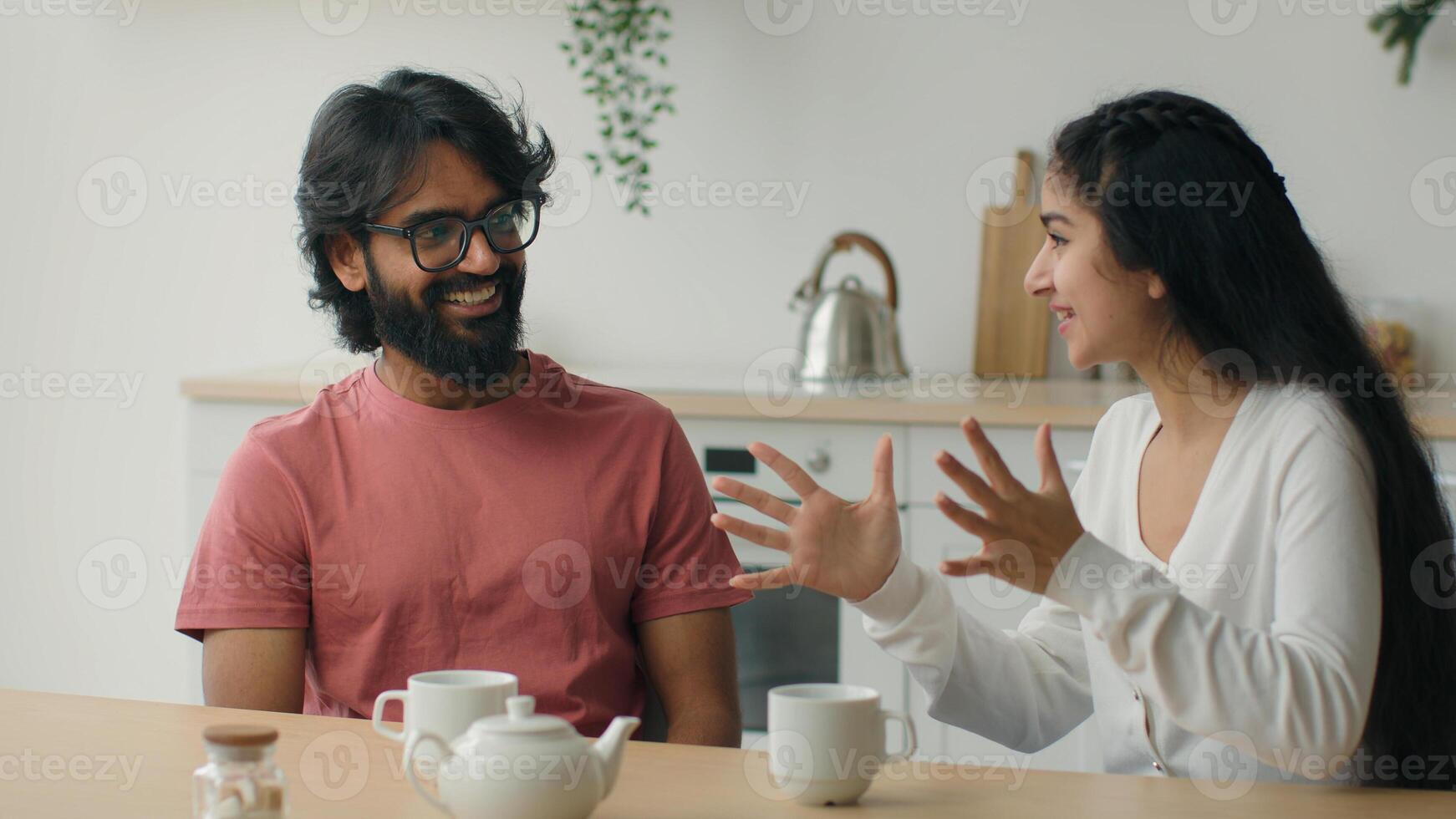 Happy multiracial couple drink coffee tea in home kitchen friendly chatter Arabian man guy listen funny story carefree conversation Indian woman girl talk storytelling romantic date friends friendship photo