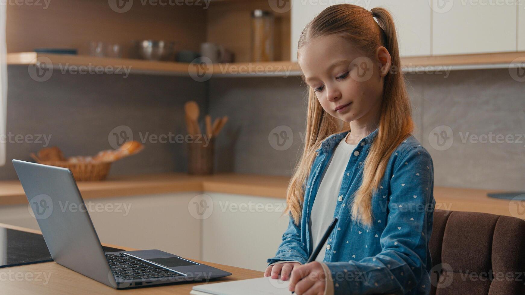 Caucasian kid girl school pupil studying online from home watching laptop web class lesson teacher by call e-learning pandemic isolation writing homework task at kitchen child remote education photo