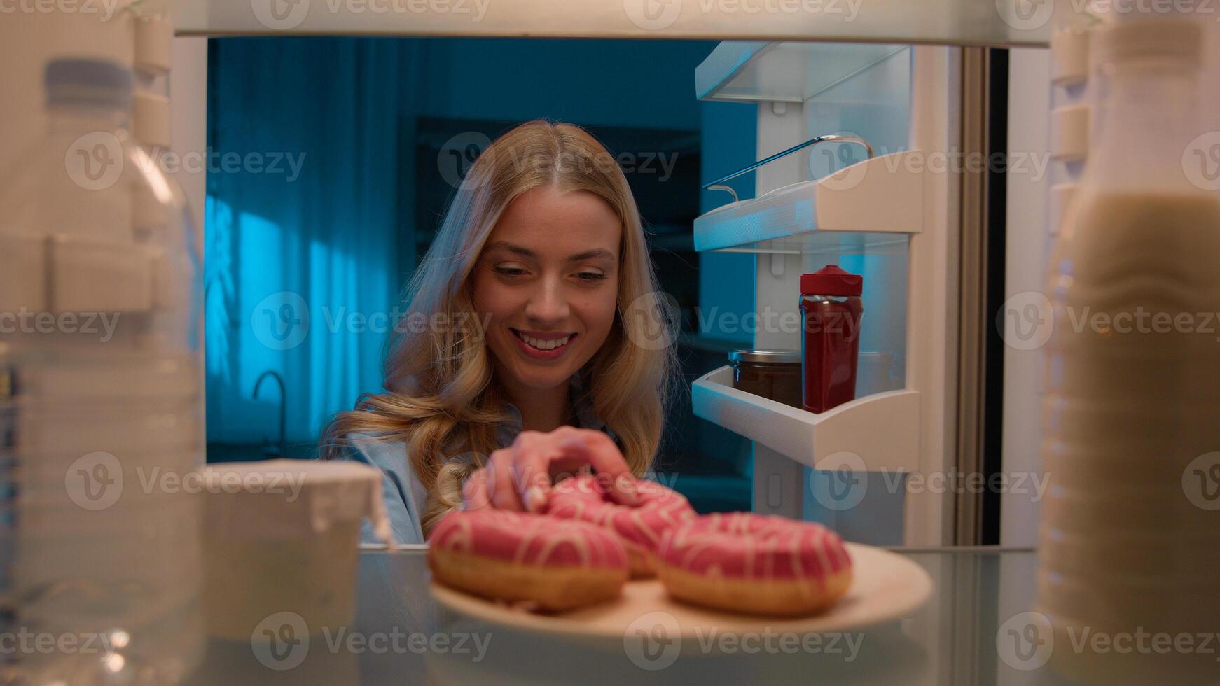 Point of view POV from inside refrigerator hungry Caucasian woman girl at night evening kitchen open fridge take out eating sweet donut unhealthy nutrition obesity diet failure overeating food addict photo