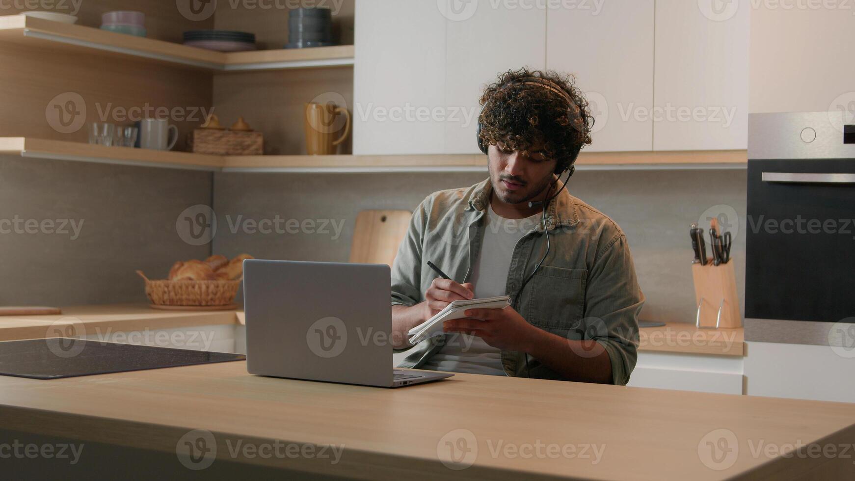 Indian man in earphones sitting at table in domestic kitchen watching educational call lecture class lesson on laptop writing notes Arabian student guy studying distant online courses quarantine photo