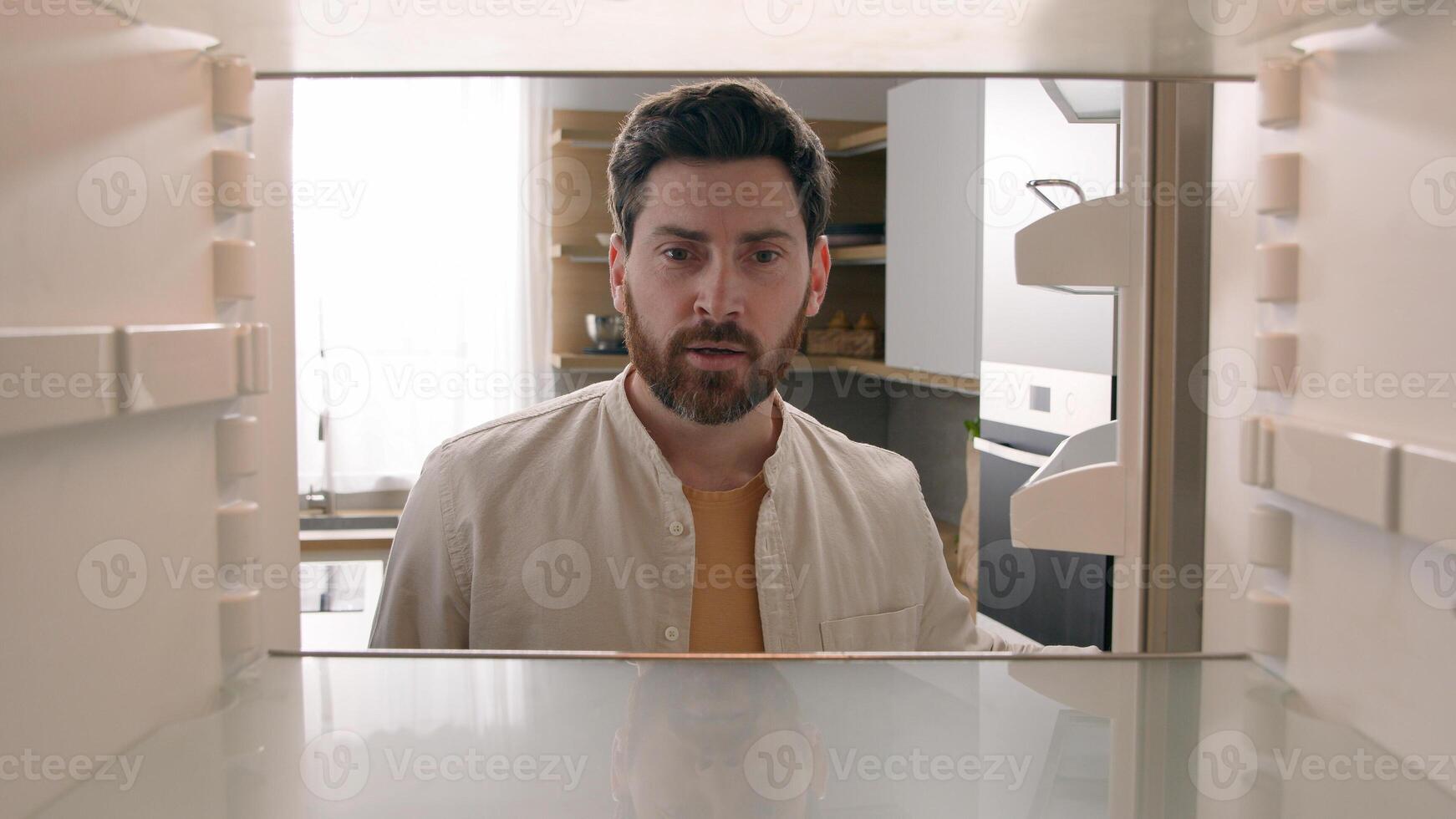 Hungry Caucasian bachelor man looking for food in empty fridge finding no meal products feeling sad disappointed poor unemployment need delivery service POV point of view from inside refrigerator photo
