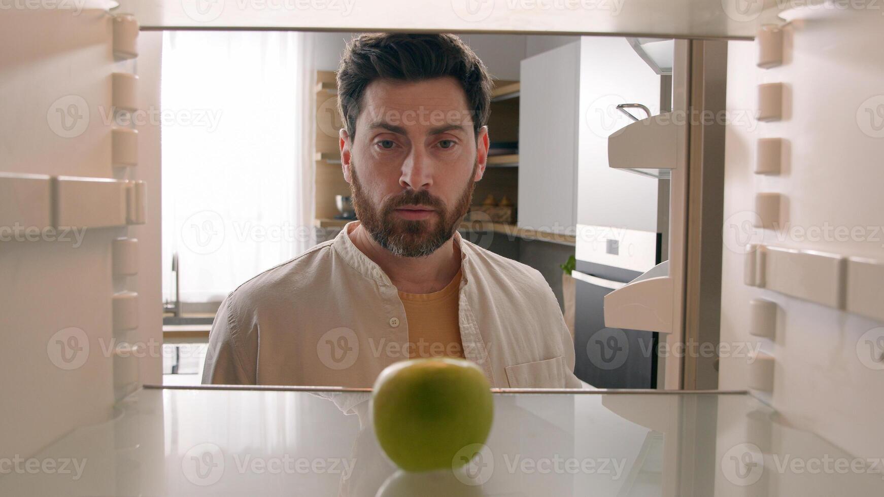 POV point of view from inside refrigerator Caucasian adult man at kitchen open empty fridge with one green apple fruit hungry guy male upset need food delivery meal crisis diet shrugs sad frustrated photo