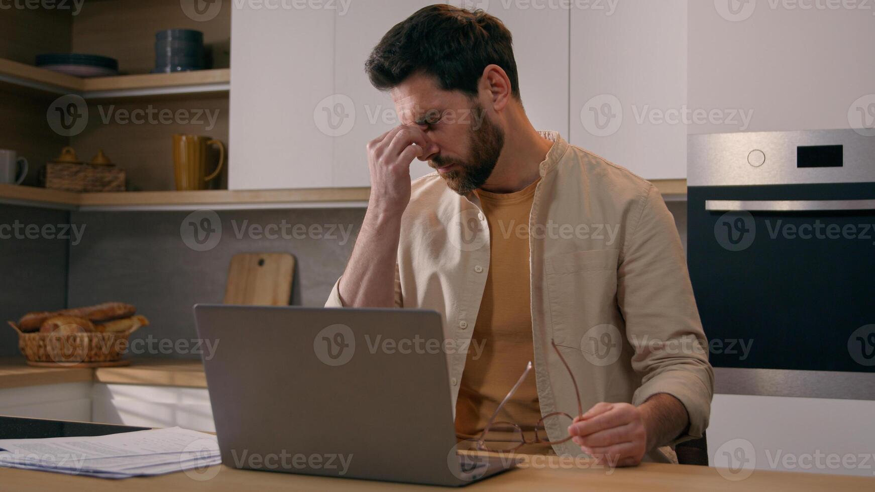 Caucasian busy freelancer man working distant from home work with laptop at kitchen exhausted businessman take off glasses suffering from eye strain pain job break from computer dry eyes overworking photo
