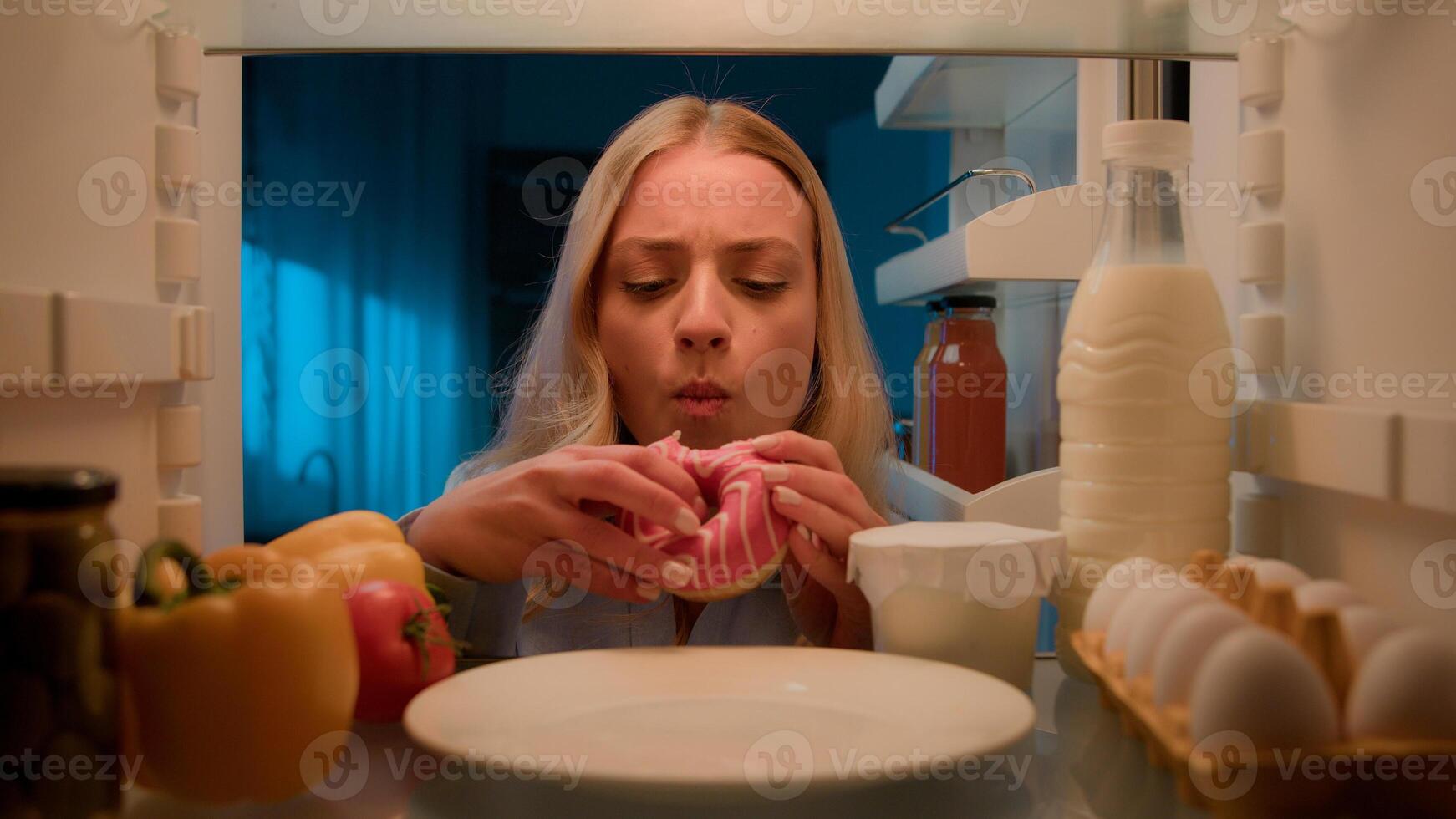 Point of view POV from inside refrigerator hungry Caucasian woman girl at night evening kitchen open fridge take out eating sweet donut unhealthy nutrition obesity diet failure overeating food addict photo