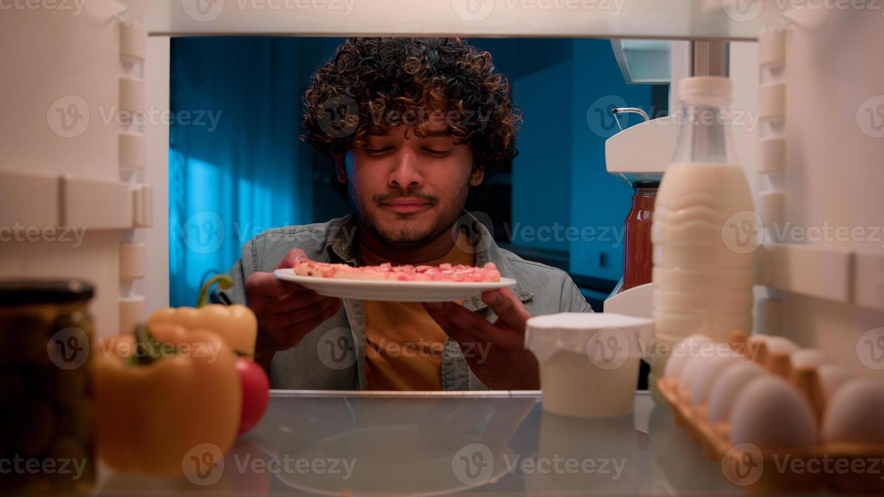 View inside from refrigerator Indian sly man with curly hair open fridge at night evening in kitchen hungry guy male looking around take out slice of pizza overeating at nighttime unhealthy nutrition photo
