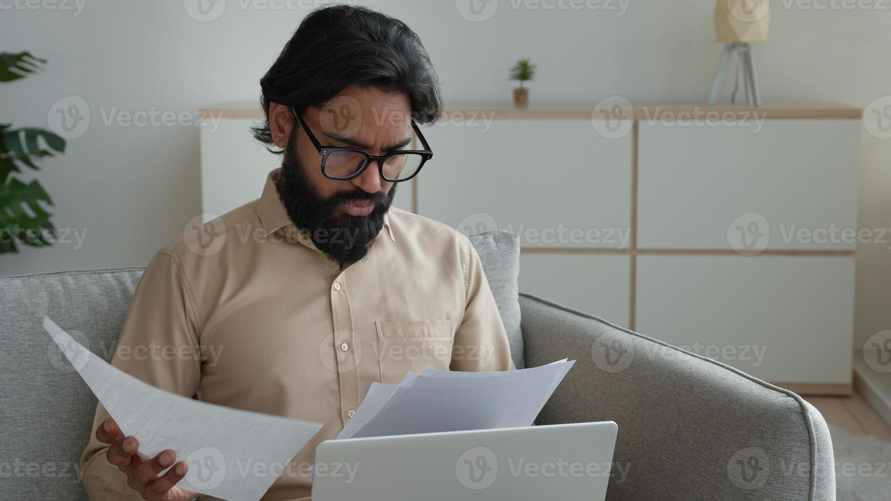 Anxious Arabian businessman freelancer on sofa reading documents pay bills at home managing finances taxes stressed man sorting out papers fees high rental rates paperwork problem budget difficulties photo