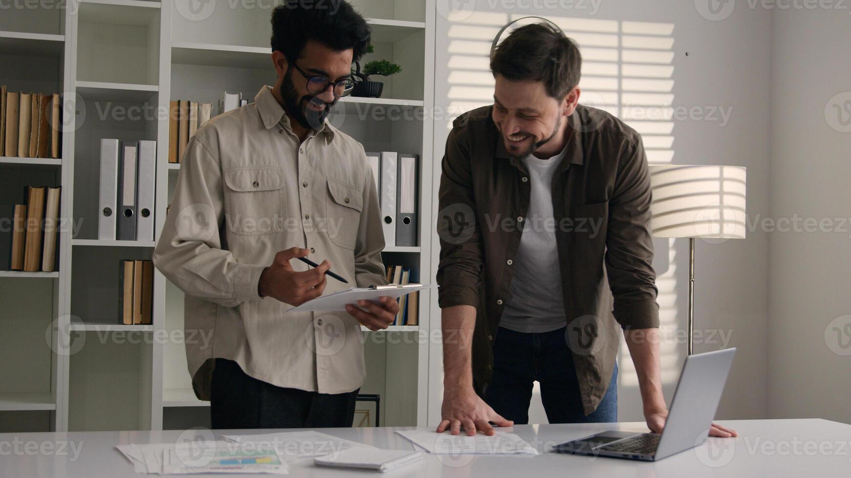 Two happy smiling multiethnic colleagues businessmen business partners at office company discuss project computer work Caucasian Indian men male coworkers look at laptop corporate teamwork discussion photo