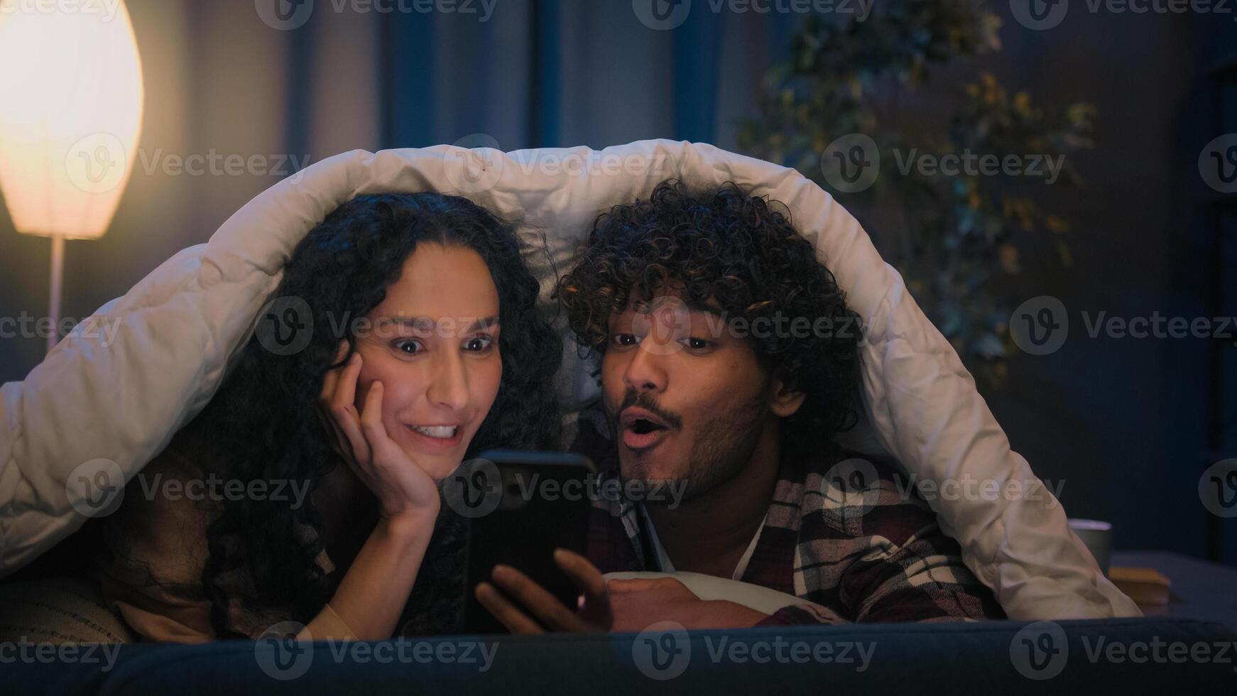 Gadget addict multiracial couple diverse man and woman scrolling mobile phone together at night under blanket social media addiction say wow amazed discount online shopping good news amazed wonder photo