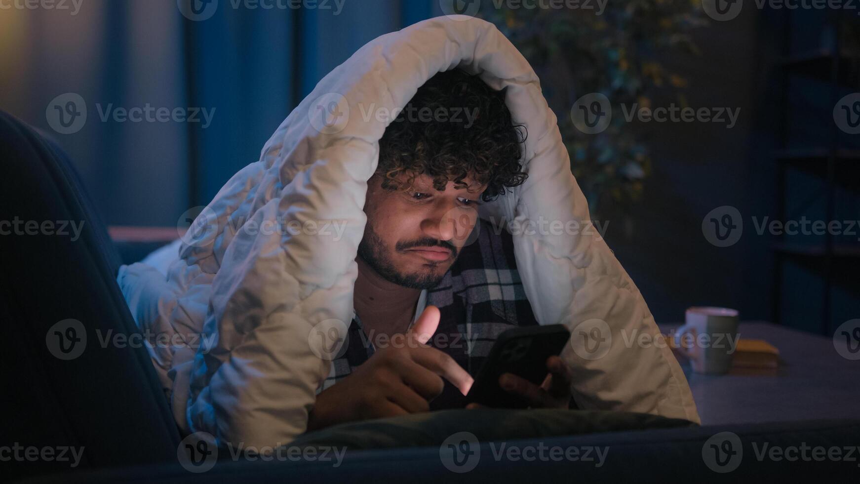 Gadget social media phone addict Indian guy Hispanic man Arabian male sleep disorder insomnia smartphone addiction at night evening home under blanket cover smiling say wow browsing cellphone news app photo