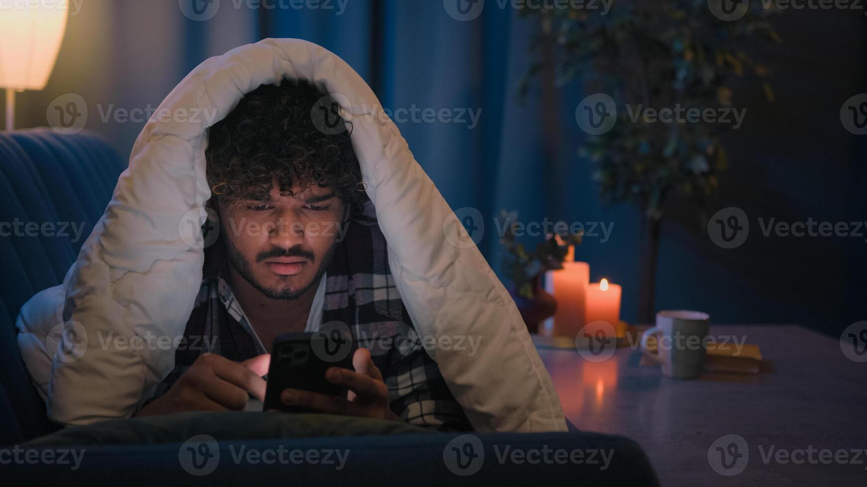 Night at home Hispanic guy gadget internet social media addict tired sleepy man reading news in social media shocked confused puzzled exhausted Arabian Indian male under blanket with mobile phone app photo