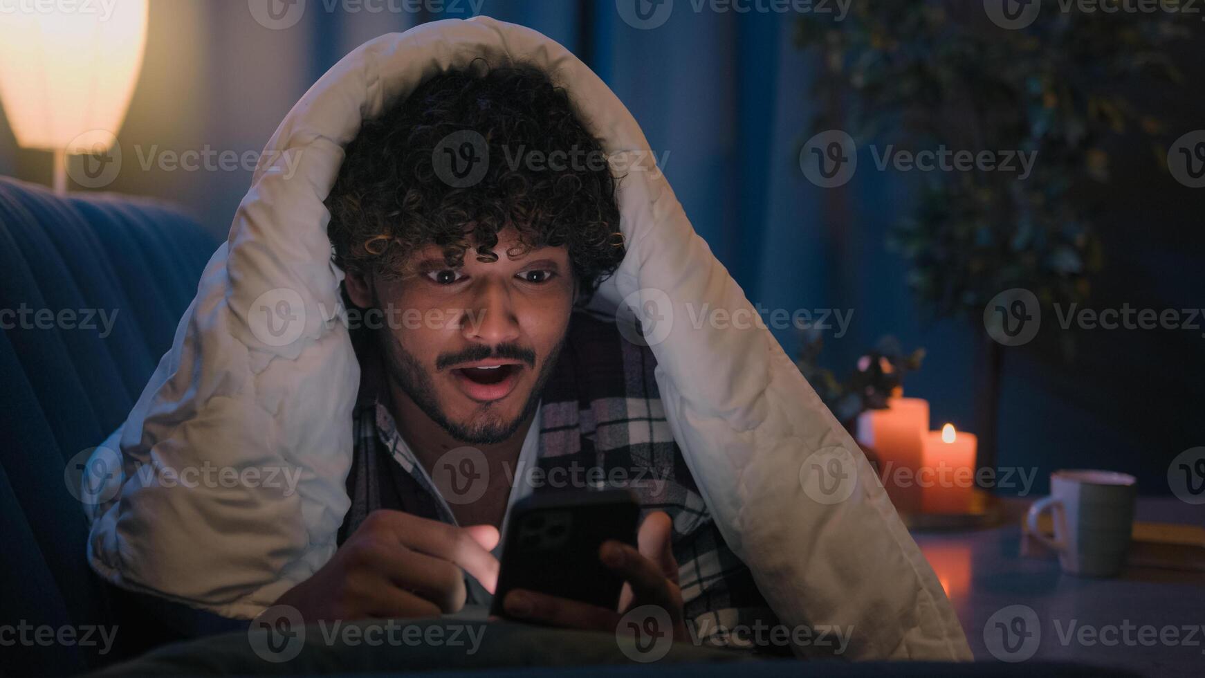 Mobile phone addict male user Indian guy surprised shocked wonder amazed reading news in smartphone at night under blanket Arabian man shock success surprise telephone victory say wow excited insomnia photo
