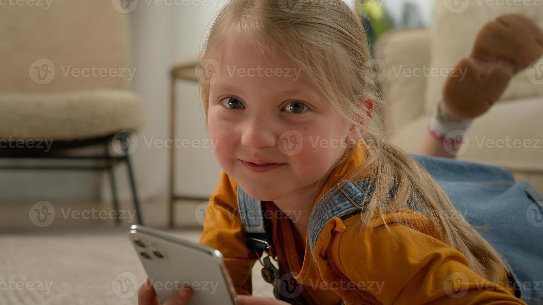 Close-up cute caucasian little kid girl lying on floor in living room using mobile phone application happy child playing telephone play online game on internet posing with smartphone at home photo