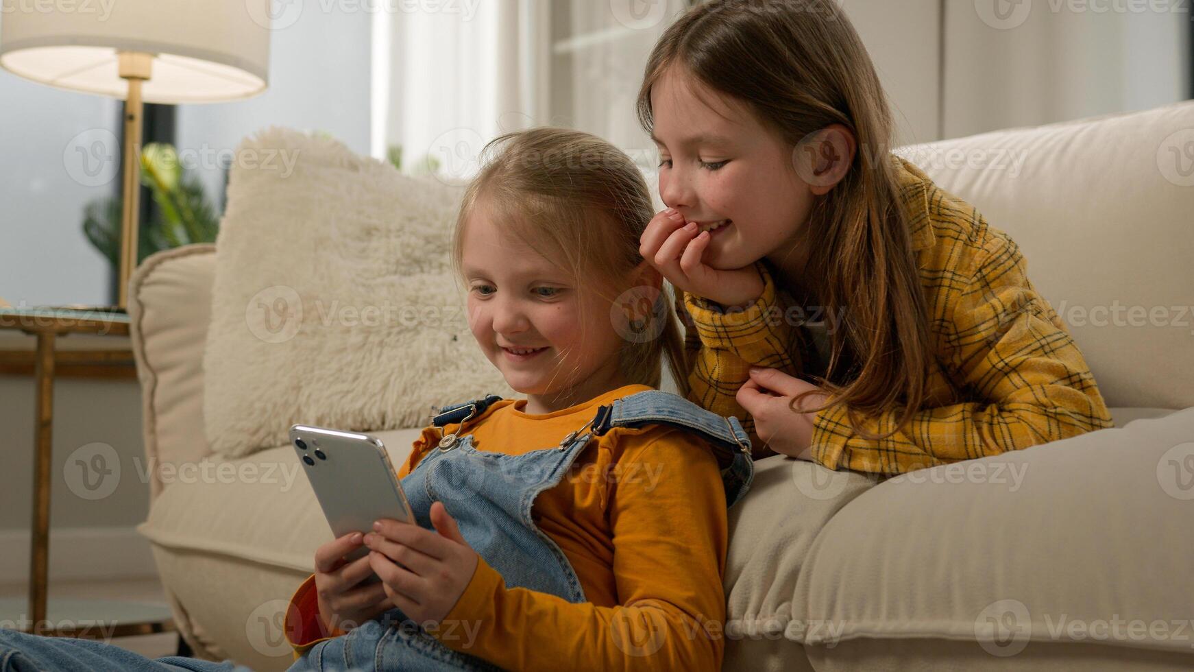 Two funny happy caucasian kids girls sisters friends children having fun playing on phone in living room couch play mobile online game together using smartphone browsing social network in Internet photo