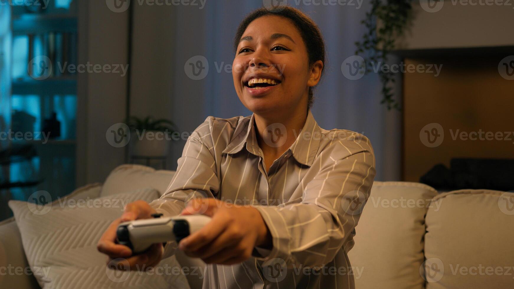 African American woman happy gamer playing console game on couch at night home smiling female use joystick play online with controller virtual cyberspace technology on smart TV in living room photo