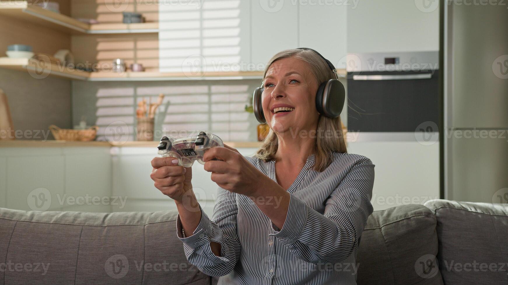 Funny smiling mature caucasian woman playing game console in headphones using joystick controller at home on couch elderly female play computer games older people and modern technology concept photo