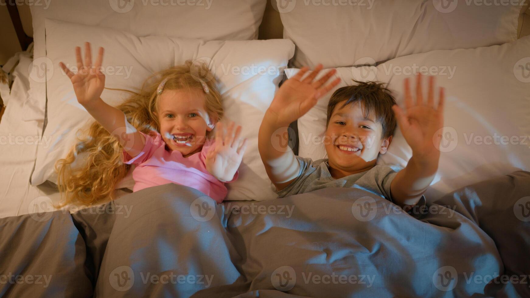 Funny playful laughing Caucasian children kids boy girl brother sister siblings happy family lying in bedroom on bed before bedtime waving hello greeting playing fooling around covered with blanket photo