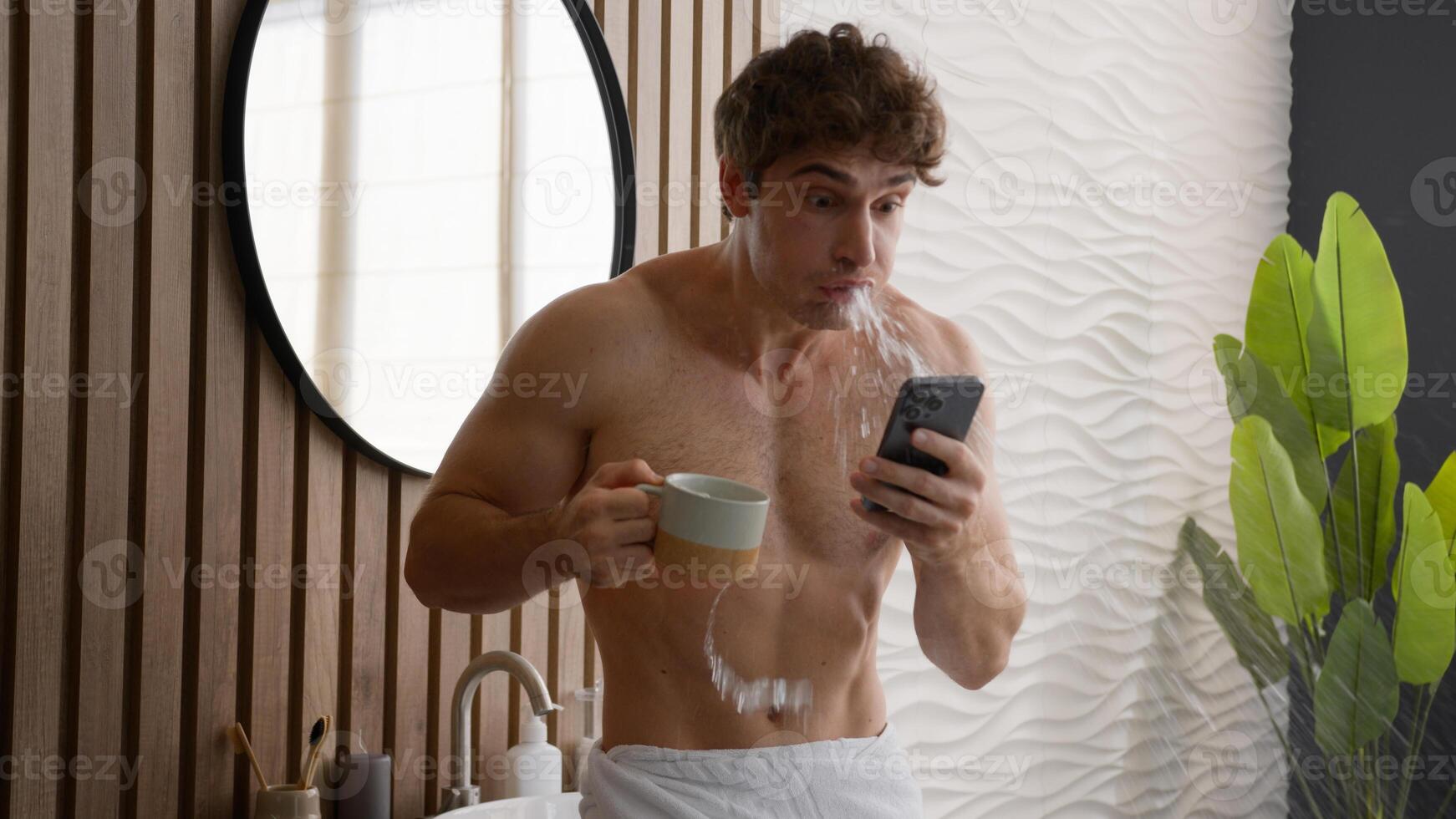 Caucasian relaxed naked man drink tea at morning routine reading news in social media in smartphone browsing at mobile phone drinking hot coffee in bathroom guy amazed shock shocked spitting beverage photo