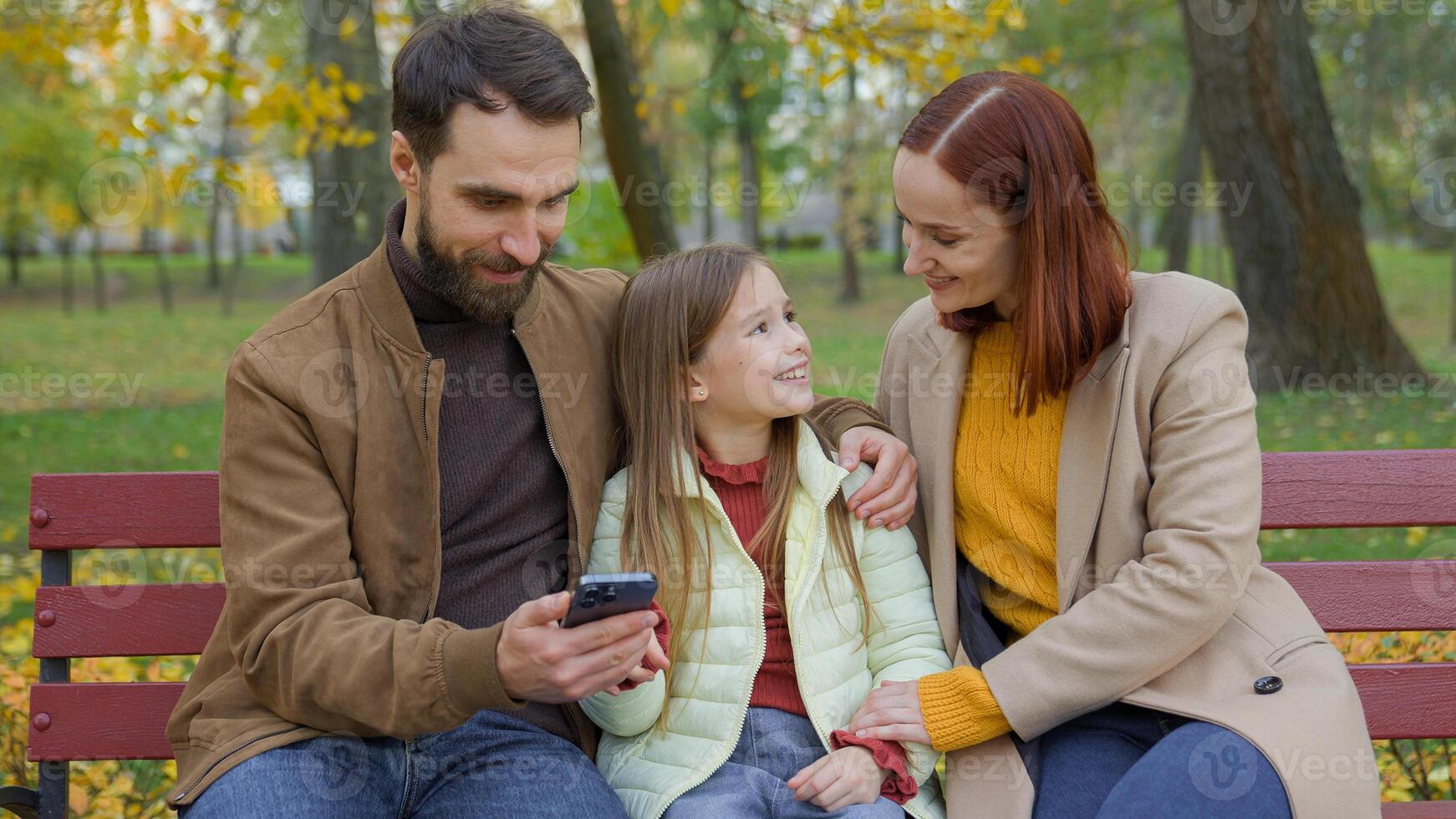 Family together happy man father woman mother child kid girl little daughter outdoors park on bench dad mom baby using mobile phone smartphone browsing choosing goods shopping online talking in city photo