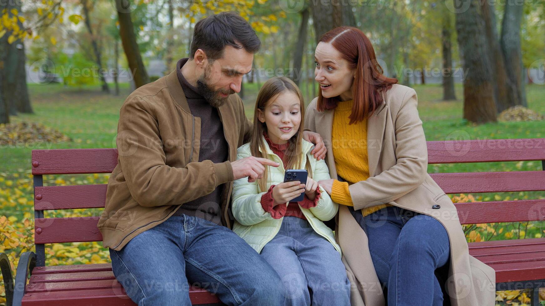 Cheerful family together happy man father woman mother child kid girl little daughter outdoors park on bench dad mom baby using mobile phone smartphone browsing amazed wonder look at screen astonish photo