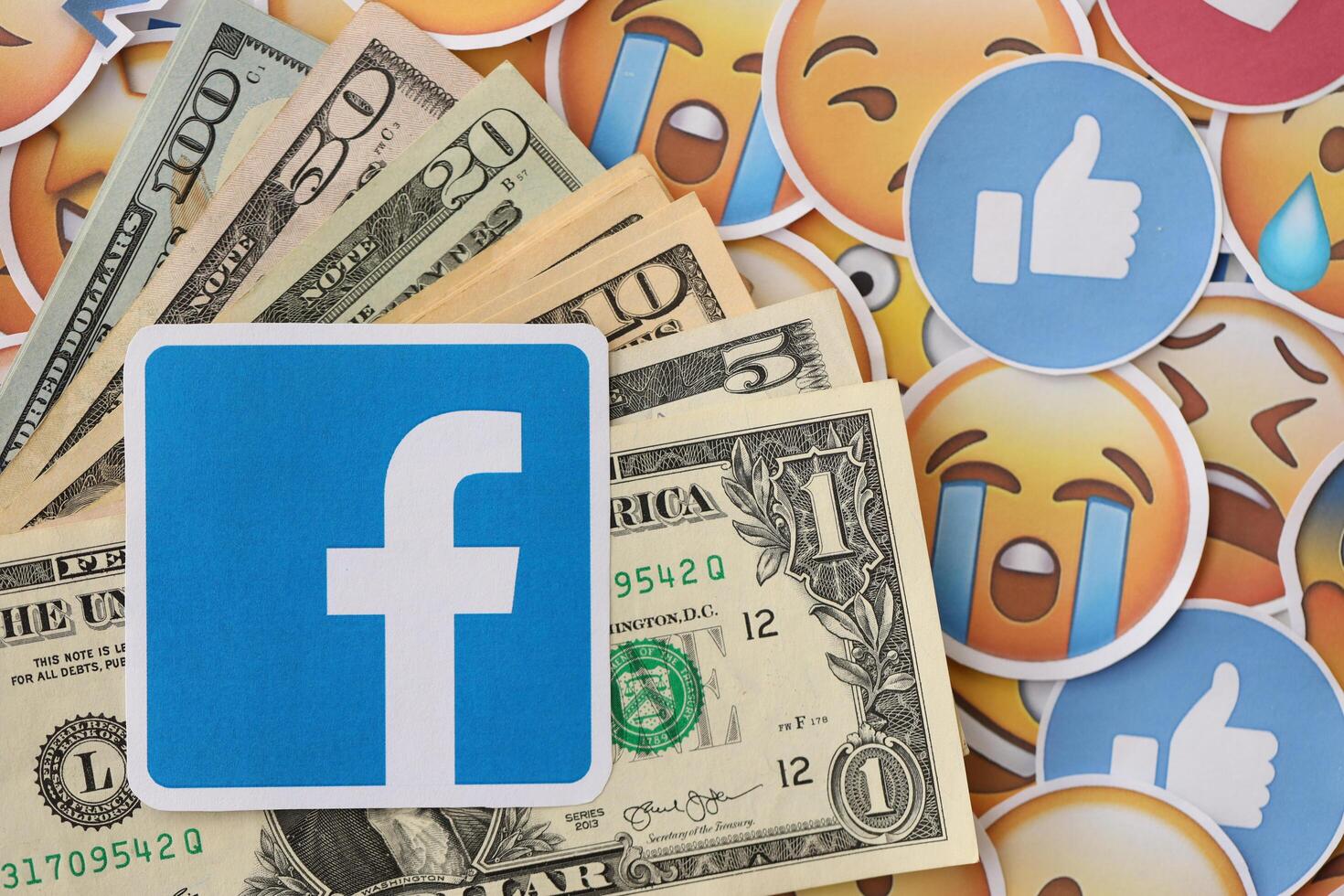 KYIV, UKRAINE - APRIL 20, 2024 Facebook logo and many usd money bills. Symbol of famous social network platform and dollar currency banknotes on lot of emoji reactions photo