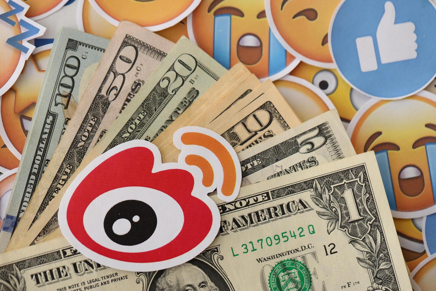 KYIV, UKRAINE - APRIL 20, 2024 Weibo logo and many usd money bills. Symbol of famous social network platform and dollar currency banknotes on lot of emoji reactions photo