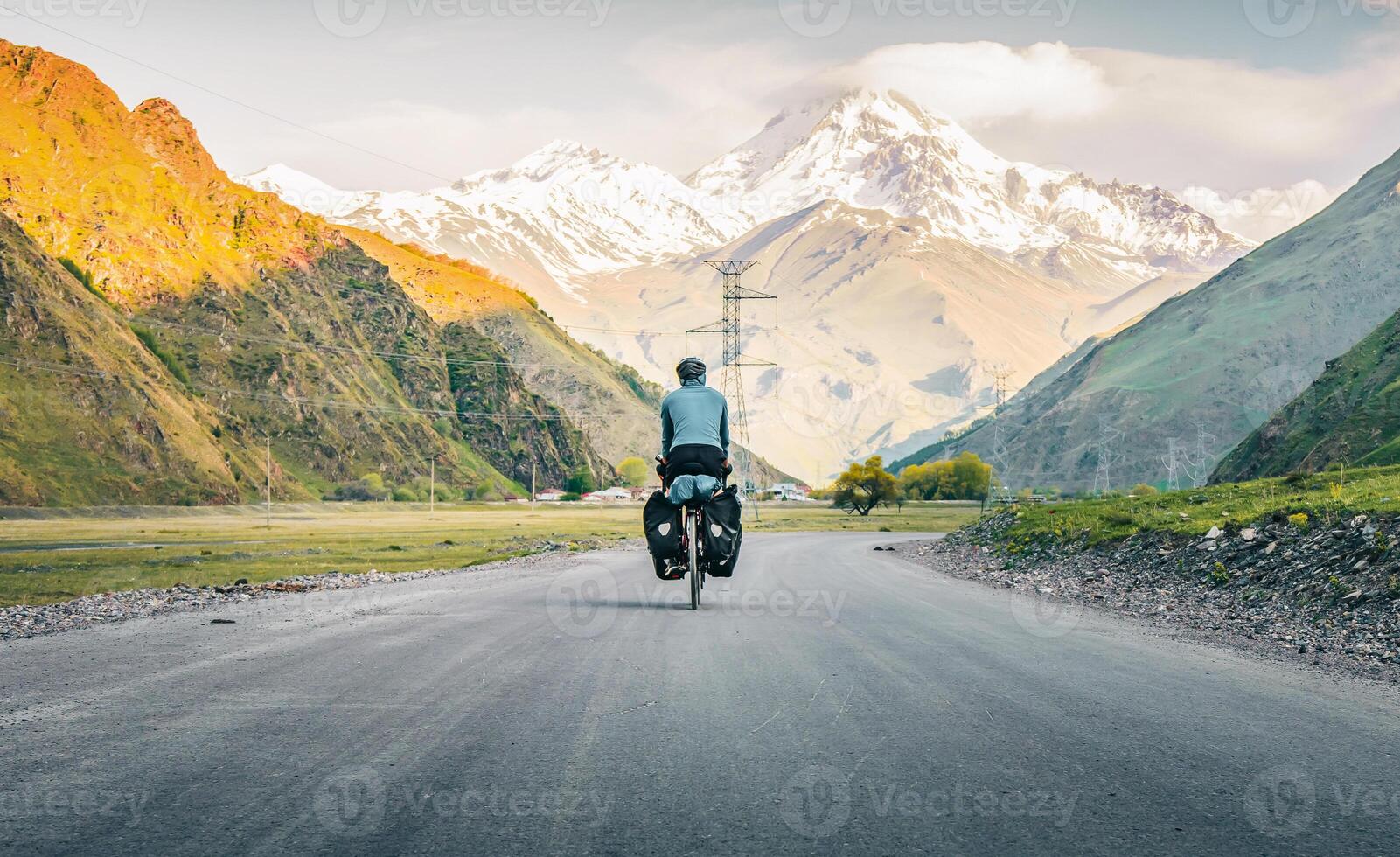 Back view one male cyclist in blue jacket pedaling on touring bicycle surrounded in mountains and green summer nature. Bicycle tour touring cycling holidays photo