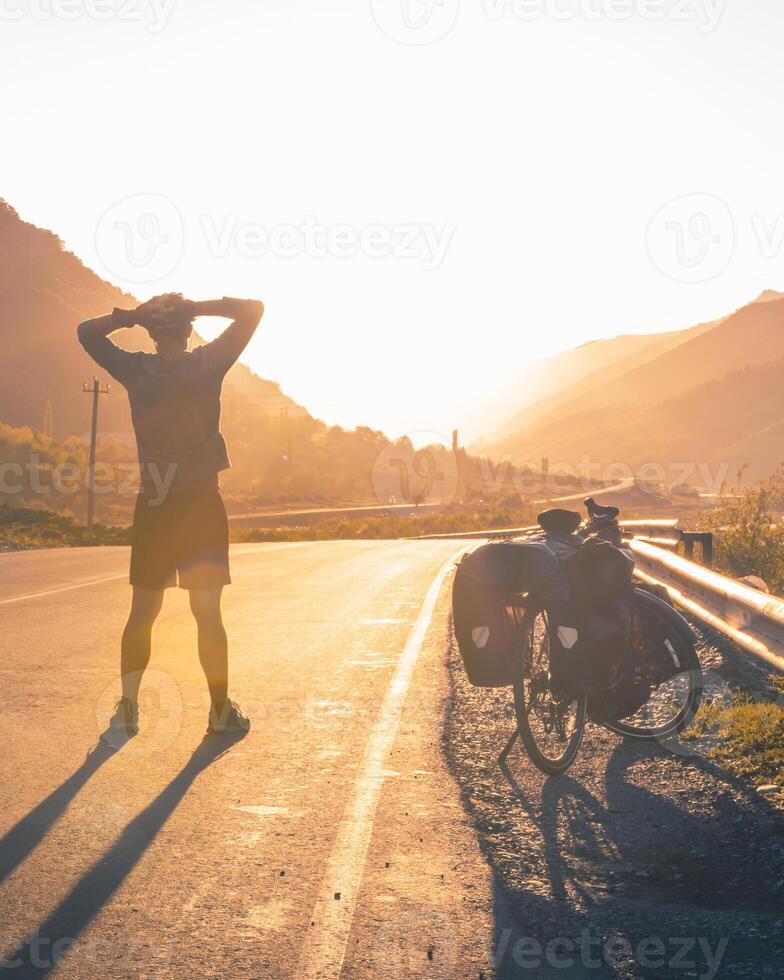 Male cyclist sportsman stands confident and determined hands on head looking to mountains by touring bicycle standing.Travel around Georgia KAzbegi national park travel destination photo