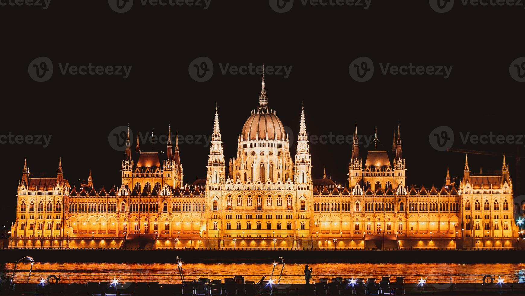 Panoramic background orange night image of Budapest parliament from the front side with boat and tourist taking a photo of the city. Travel in hungary and historical sites.