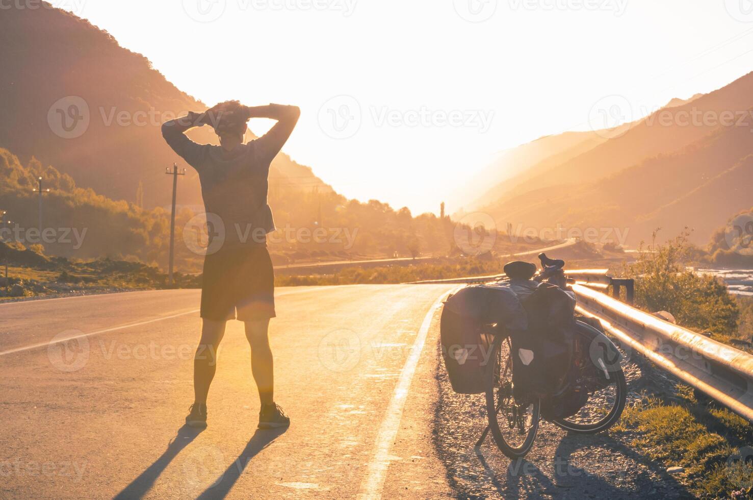 Male fit cyclist stand by touring bicycle enjoy sunset with caucasus mountains background. Cycling holidays and eco-friendly, sustainable healthy travel concept photo