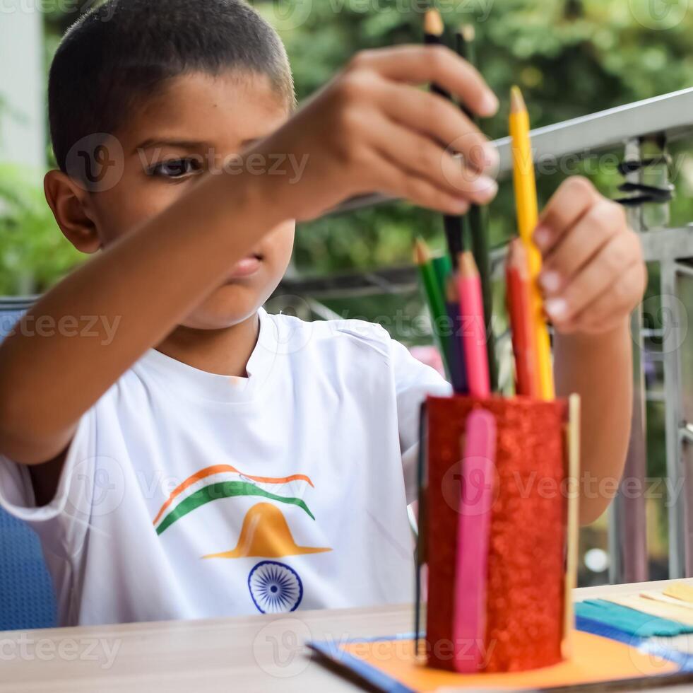 Smart Indian little boy performing best out of waste activity with different colourful objects during the summer vacations, Cute Indian Kid doing best our of waste activity at home on wooden table photo
