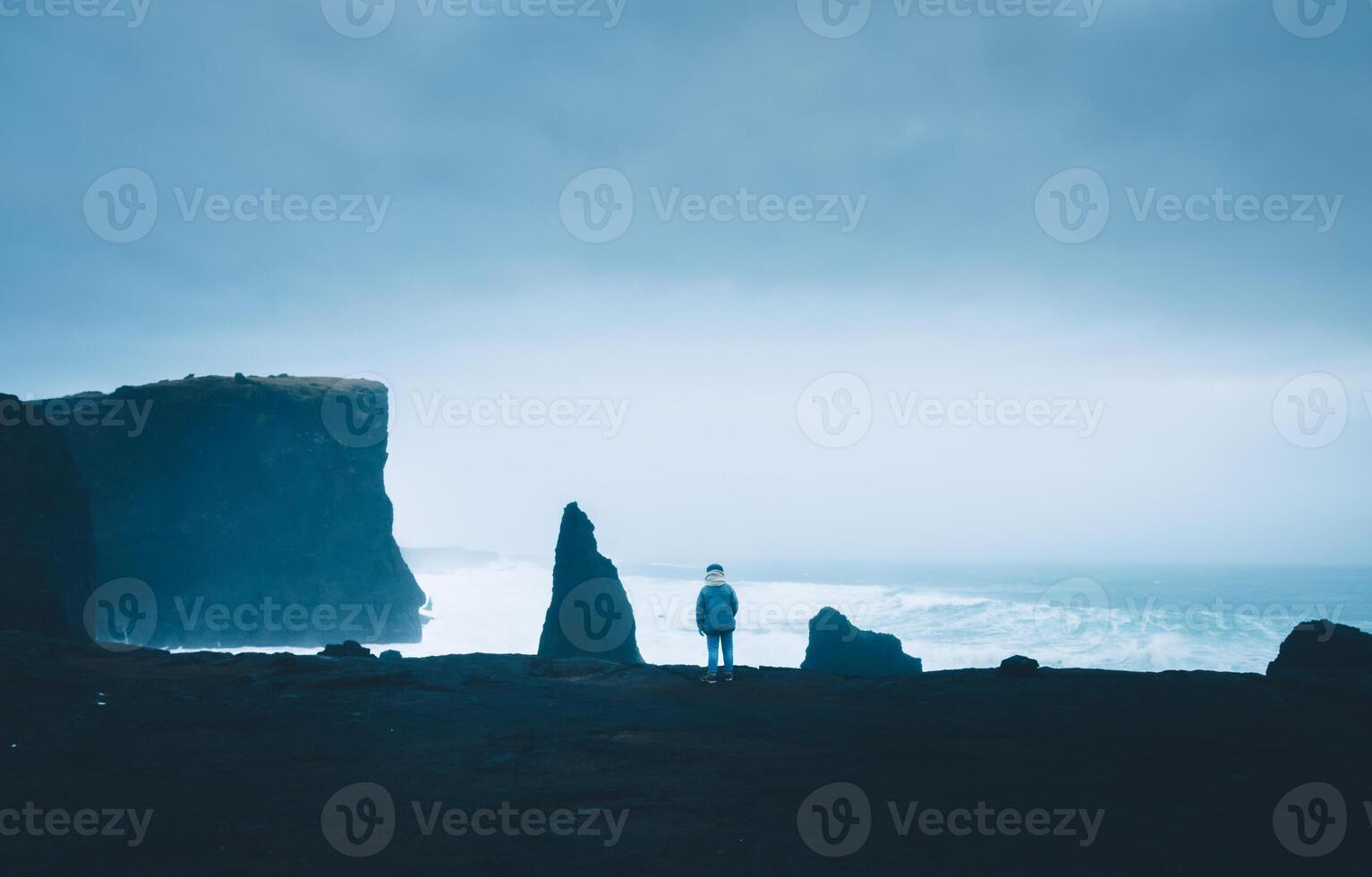 Woman tourist stand in very windy day watch Atlantic ocean waves crash to rocks in Reykjanes peninsula shore in Iceland. Popular scenic nature location. Iconic Valahnukamol stack photo