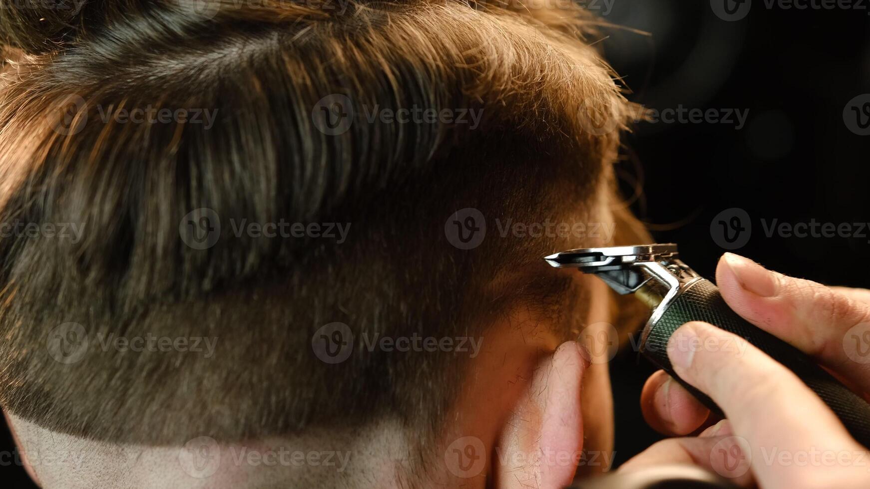 close up Shot of a handsome barber giving a haircut to his client using trimmer. Hairdresser service in a modern barbershop in a dark key lightning with warm light back view photo