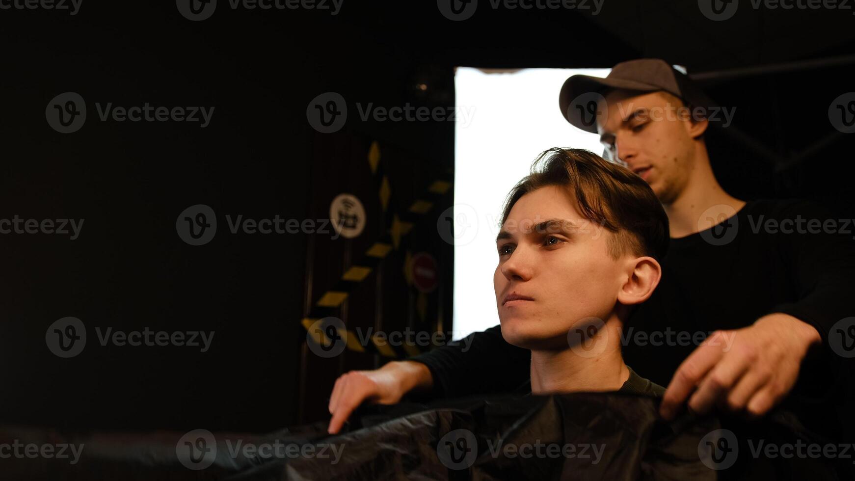 Caucasian man in shirt sitting on the chair while barber closing his client with black peignoir in salon. Hairdresser service in a modern barbershop in a dark key lightning with warm light side view photo