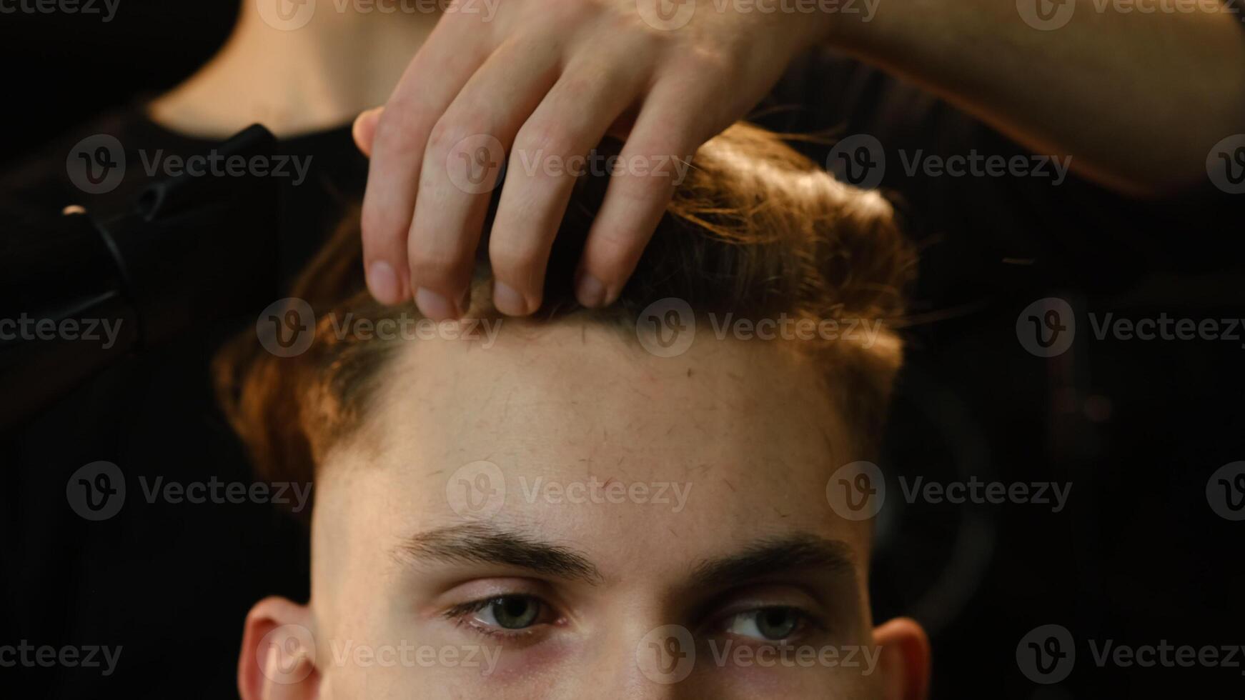 Close up young barber drying hair of customer with hair dryer in barber shop in a dark key lightning with warm light and correct hair with hand. Hairdresser for man front view photo