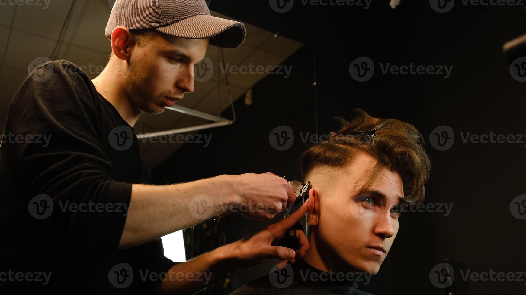 men's hairstyling and haircutting with hair clipper in a barber shop or hair salon. Hairdresser service in a modern barbershop in a dark key lightning with warm light side view photo