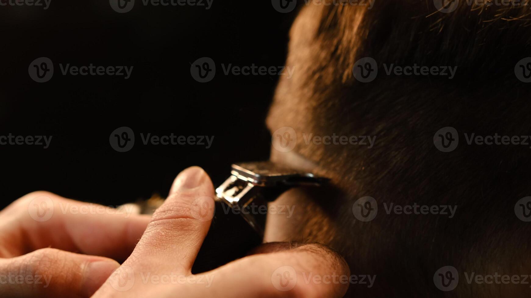 close up Shot of a handsome barber giving a haircut to his client using trimmer. Hairdresser service in a modern barbershop in a dark key lightning with warm light back view photo